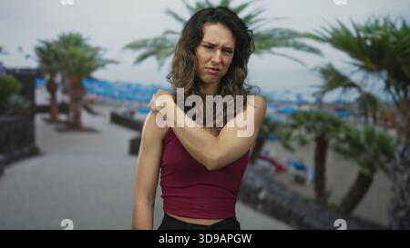 Frau in ärmellosem Oberteil, die Schulter neben Palmen auf der Strandstraße hält, während sie von Muskelanstrengung zuzwinkert; Schmerzen. Stockfoto