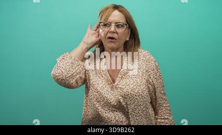 Frau, die Hand an Ohr schröpft und hinter einer Brille in einem Studio-blaugrünen Hintergrund knirscht; Hörschwierigkeiten sind besorgniserregend. Stockfoto