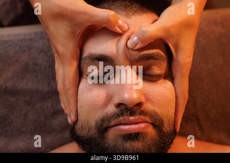 Männliche Schönheit - Mann erhält Gesichtsmassage im Luxus-Spa. Gutaussehender Kerl, Gesichtsmassage. Hände einer Masseurin. Gutaussehender Mann im Spa, der sich einen F.C. Stockfoto