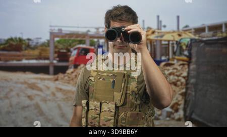 Mann Soldat in Tarnweste hält ein Fernglas an die Augen und reibt sich dann seine Stirn, während er in der Nähe einer Baustelle steht; Wachsamkeit bei Ermüdung Stockfoto
