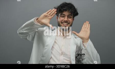 Junger hispanischer Mann mit Bart in Brille Lächeln und Gesten in einem weißen Mantel vor grauem Hintergrund, der Selbstvertrauen und Freundlichkeit zeigt. Stockfoto