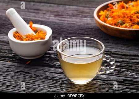 Tasse Calendula Tee, begleitet von getrockneten Blütenblättern und einem weißen Mörtel mit Calendula Blumen - Calendula officinalis Stockfoto