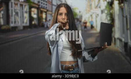 Die junge hispanische Frau zeigt mit dem Finger auf die Kamera, während sie einen Laptop hält und auf einer belebten Straße ihre Nase kneift; ekelhafte Vorsicht. Stockfoto