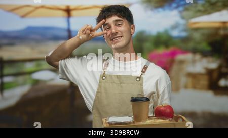 Mann mit Schürze, der Holztablett mit Kaffeetasse, Gebäck und Apfel auf der Terrasse des Restaurants hält, die Hand zeigt ein Friedenszeichen vor Augen; freundlicher Service. Stockfoto