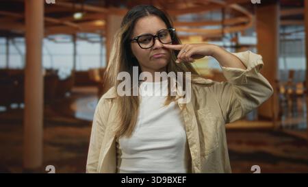 Frau Finger an Nase im Restaurant mit Brille und beigem Hemd, geschlossenen Augen und leichtem Lächeln; verspieltes Selbstvertrauen. Stockfoto