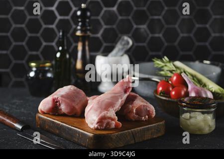 Zwei rohe Hasenbeine und Sattel auf einem hölzernen Schneidebrett, umgeben von Kochzutaten wie Kirschtomaten, Kräutern und Gewürzen Stockfoto