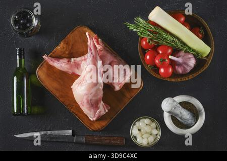 Blick von oben auf zwei rohe Hasenbeine auf einem Holzbrett, umgeben von frischem Gemüse, Kräutern und Gewürzen. Die Szene erfasst die Zutaten für einen Stockfoto