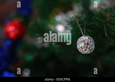 Festliche Nahaufnahme mit glänzenden silbernen Disco-Ball-Ornamenten, die an grünem Weihnachtsbaumzweig hängen. Verschwommener Hintergrund mit roter Kugel sorgt für einen fröhlichen Urlaub Stockfoto