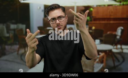 Junger Mann mit Bart, der auf der Terrasse des Stadtcafés unhöfliche Gesten macht, umgeben von einem Restaurant im Freien mit Tischen und Stühlen. Stockfoto