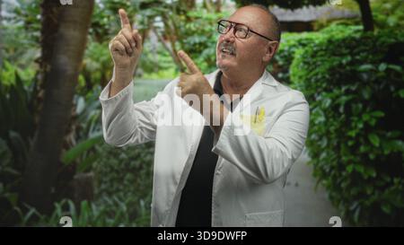 Mann Doktor, Senior hispanic im Laborkitt zeigt mit beiden Händen den Finger nach oben in die Nähe des Gartenturms am Gebäude, trägt Brille und Taschenstifte; Kuriosit Stockfoto