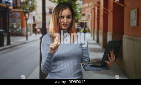 Frau, die einen Laptop hält, zeigt mit dem Zeigefinger auf eine Fußgängerzone, die von Bögen und Geschäften gesäumt ist, rote Haare und lässiger Pullover sichtbar; Selbstvertrauen. Stockfoto