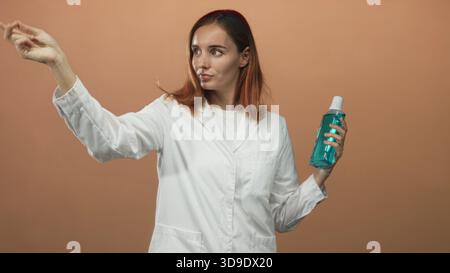 Frau Ärztin und junge Rothaarige im weißen Labormantel hält eine blaue Flasche mit Kappe und zeigt den Zeigefinger in Richtung des Betrachters im Studio; Hygieneanleitung Stockfoto