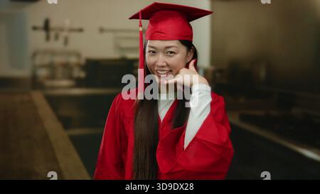 Frau in roter Abschlusskleidung steht lächelnd in einer Küche und macht eine Handygeste, die Leistung in einer Inneneinrichtung verkörpert. Stockfoto