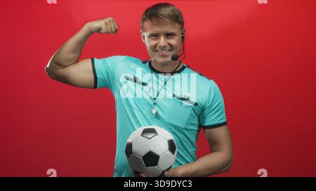 Der Schiedsrichter hält Fußball, trägt Pfeife und Headset, beugt den Bizeps und posiert vor rotem Studio-Hintergrund; Selbstvertrauen. Stockfoto