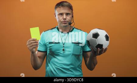 Mann Schiedsrichter hält gelbe Karte und Fußball, Hand hochgehoben mit Pfeife sichtbar im Studio; Autoritätsentscheidung. Stockfoto