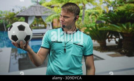 Mann Schiedsrichter hält Fußball in der rechten Hand mit Pfeife und Headset am Poolgebäude; Entscheidungsbefugnis. Stockfoto