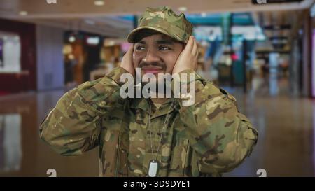 Hispanischer Mann in Tarnuniform bedeckt die Ohren mit beiden Händen inmitten des überfüllten Einkaufszentrums; Unwohlsein. Stockfoto