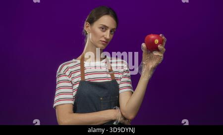 Frau in gestreiftem Hemd und Denim-Schürze mit rotem Apfel mit Handschuhen in lila Atelier; Hygieneinspektion. Stockfoto