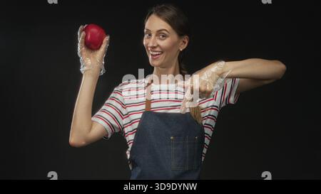 Frau, die roten Apfel hält und mit Handschuhen im Studio vor schwarzem Hintergrund zeigt; gesunde Snackfreude. Stockfoto