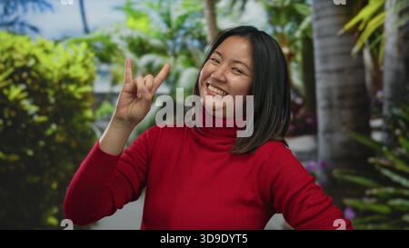 Junge Frau in rotem Pullover, die Gesten macht und in einem lebhaften grünen Park im Freien weit lächelt, der Spaß und fröhliche Energie vermittelt. Stockfoto