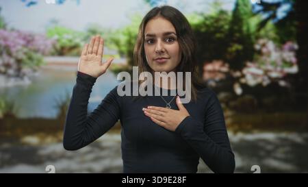 Die Frau hebt die rechte Hand und legt die linke Hand über ihr Herz in einem Wald an einem Teich mit blühenden Sträuchern und grünem Laub; Ehre. Stockfoto