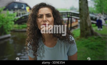 Frau runzelt mit gefurchten Augenbrauen im grünen Park an einer Holzbrücke über einem Teich unter weichem Tageslicht; ruhige Introspektion. Stockfoto