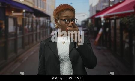 Frau mit Brille und goldenen Ohrringen in schwarzem Blazer zeigt mit dem Finger auf den Tempel auf der Straße; nachdenkliche Introspektion. Stockfoto