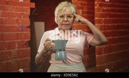 Eine Frau, die einen Mokakopf hält, zeigt mit dem Finger auf den Tempel im Studio mit freiliegender Ziegelwand, trägt Brille und rosa Polo; nachdenkliche Reflexion. Stockfoto