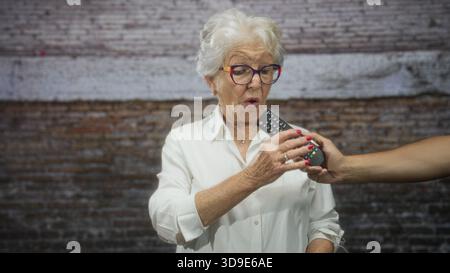 Ältere Frau in weißer Bluse zeigt eine fernseh-Fernbedienung in Richtung Kamera mit Heckgesicht und verlängertem Arm im Studio; Unabhängigkeitsbelästigung. Stockfoto