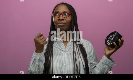 Frau mit Brille, die Uhr vor rosa Hintergrund hält und Zeitmanagement-Konzept vermittelt. Stockfoto