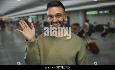 Junger Mann, der am Flughafenterminal lächelt und mit einer Welle in geschäftiger Umgebung begrüßt, die Reisen und Multikulturalität in der modernen Transitlounge mit zeigt Stockfoto
