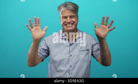 Mann mit grauen Haaren lächelt und winkt mit offenen Händen im Studio vor türkisfarbenem Hintergrund; warmer, fröhlicher Gruß. Stockfoto