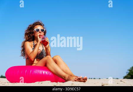 Wunderschöne Frau mit lockigen Haaren, die sich am tropischen Strand mit Erfrischungsgetränk und rosa Schwimmring entspannt. Stockfoto