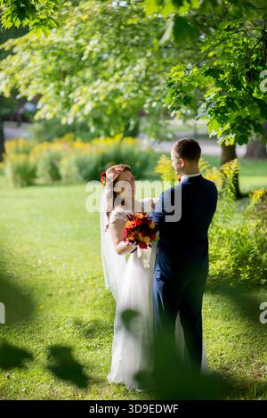 Braut und Bräutigam stehen umarmt unter einem üppig grünen Baumdach, Braut lächelt den Bräutigam herzlich an und hält einen hellen Blumenstrauß – ein zartes und natürliches Wir Stockfoto