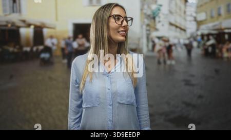 Frau lächelt mit Brille und blauem Hemd, während sie auf dem straßenplatz mit verschwommener Menschenmenge steht; Selbstvertrauen reist. Stockfoto