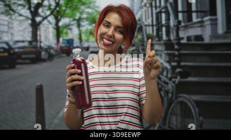 Frau mit rotem Haar hält eine Flasche Saft und zeigt mit dem Zeigefinger auf die Straße neben dem Fahrrad und Steintreppen; fröhliches Wellness. Stockfoto