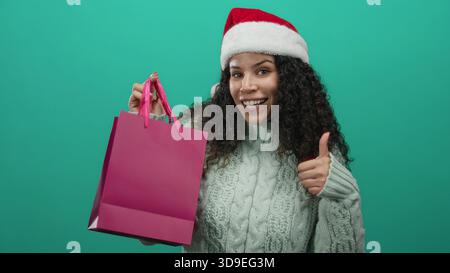 Glückliche junge hispanische Frau mit weihnachtsmann-Hut und lockigem Haar gibt eine Daumen-nach-oben-Geste, während sie eine rosa Einkaufstasche gegen einen grünen isolierten Backgr hält Stockfoto