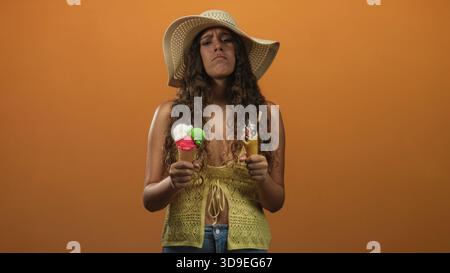 Frau, die zwei Eiskegel hält und die Stirn runzelt, während sie in einem Studio schmelzende Schaufeln vor einem orangen Hintergrund vergleicht; Enttäuschung der Unentschlossenheit im Sommer Stockfoto