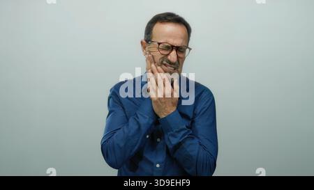 Hispanischer Mann, der Zahnschmerzen vor weißem Hintergrund erlebt, seinen Kiefer mit einem schmerzhaften Ausdruck umklammert, isoliert in einem älteren Reifen Kontext Showi Stockfoto