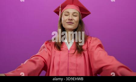 Eine junge Frau in Abschlusskleidung lächelt selbstbewusst vor einem leuchtenden rosa Hintergrund und verkörpert ein Gefühl von Leistung und Freude. Stockfoto