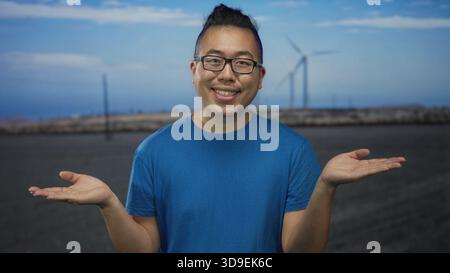 Mann mit den Handflächen auf der Straße, mit blauem T-Shirt und Brille, stehend vor Windmühlenfeld und Himmel; Neugier. Stockfoto