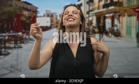 Frau, die eine Kreditkarte hält und mit dem Finger auf die Brust zeigt, während sie auf der Straße lächelt und eine Brille trägt; Selbstbewusstsein. Stockfoto