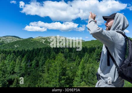 Ein Mann mit Outdoor-Kleidung und Rucksack genießt an einem hellen Sommertag eine malerische Berglandschaft. Er steht über einem üppig grünen Wald und sieht aus Stockfoto