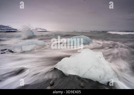 Blick auf Gletschereisbrocken, die auf schwarzem Sand ruhen, während die Wellen unter einem bewölkten Himmel um sie herum stürzen, Sveitarfelagid Hornafjordur, Sveitarfelagid Hornafjor Stockfoto