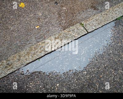 Reflektierende Wasserpfütze auf einer dunklen, strukturierten Asphaltoberfläche neben einer verwitterten Betonkante Stockfoto