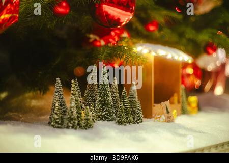 Miniatur-Bahnhof im Weihnachtsdorf mit kleinen Kiefern und Schnee unter einem großen geschmückten Baum. Nahaufnahme, geringe Schärfentiefe, kein p Stockfoto