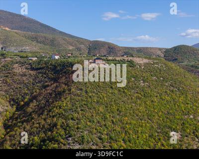 Aus der Vogelperspektive auf einen grünen Bergkamm in der Nähe von Kukës, mit verstreuten Häusern auf dem Gipfel unter einem klaren blauen Herbsthimmel. Stockfoto