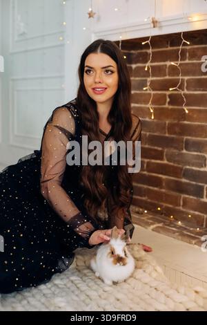 Eine junge Frau mit langen, welligen Haaren trägt ein schwarzes Kleid mit Glitzern und sitzt auf einem gemütlichen Teppich neben einem gemauerten Kamin. Stockfoto