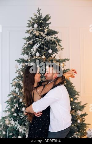 Ein fröhliches Paar umarmt sich vor einem geschmückten Weihnachtsbaum. Das Zimmer hat eine warme Atmosphäre, voller Weihnachtsdekoration und dem Geist von Cell Stockfoto