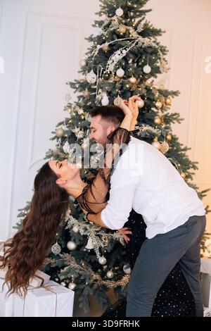 Ein Paar genießt einen lustigen und verspielten Moment zusammen um einen wunderschön geschmückten Weihnachtsbaum, umgeben von umhüllten Geschenken, die einen fröhlichen Urlaub schaffen Stockfoto